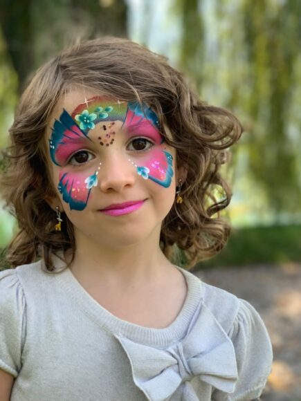 Portrait eines dunkelhaarigen Kindes an einem Kindergeburtstag, mit professioneller Gesichtsbemalung in den Farben lila und blau. Das Sujet erinnert an einen Schmetterling.
